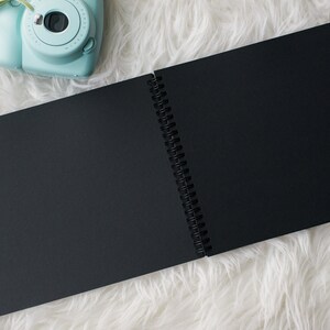 May include: A black spiral bound notebook with blank pages, open to reveal the white binding. The notebook is lying on a white fluffy surface. A light blue instant camera with a lens is visible in the top left corner of the image.