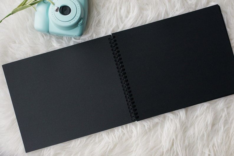 May include: A black spiral bound notebook with blank pages, open to reveal the white binding. The notebook is lying on a white fluffy surface. A light blue instant camera with a lens is visible in the top left corner of the image.