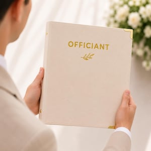 May include: A cream-colored binder with gold-toned metal corner protectors and a gold-stamped cover featuring the word "OFFICIANT" above a small leaf icon. The binder has a smooth, matte finish and a vertical spine with visible gold-toned rivets.