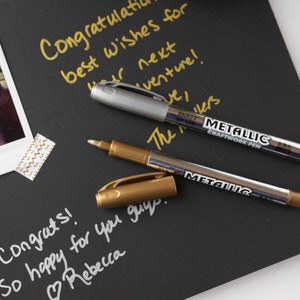 May include: Two metallic markers, one gold and one silver, on a black surface. The markers are labeled "Metallic Craftwork Pen". The text "Congrats! So happy for you guys. Rebecca" is written in white on the surface.