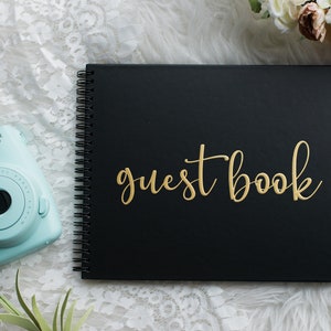 May include: Black guest book with gold lettering that says "guest book". The book is laying on a white fuzzy surface with a light blue instant camera and a bouquet of pink flowers.