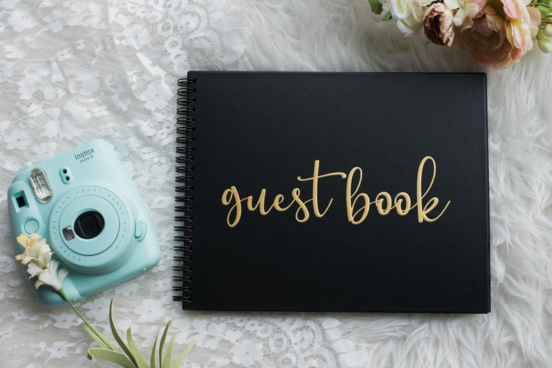 May include: Black guest book with gold lettering that says "guest book". The book is laying on a white fuzzy surface with a light blue instant camera and a bouquet of pink flowers.