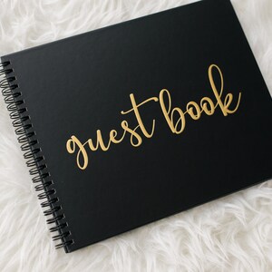May include: Black guest book with gold lettering that reads "guest book". The book is closed and lying on a white fluffy surface.