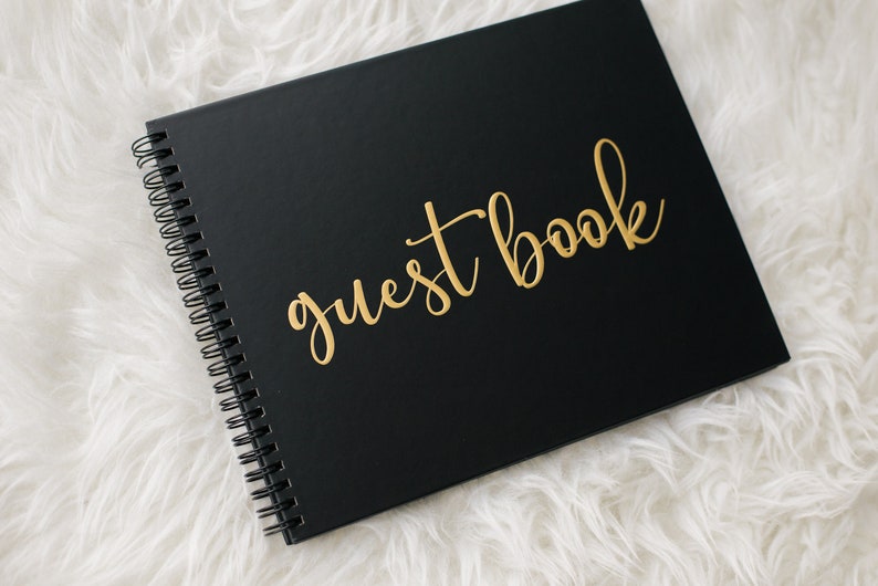 May include: Black guest book with gold lettering that reads "guest book". The book is closed and lying on a white fluffy surface.