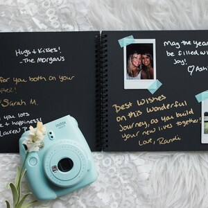 May include: A black scrapbook with gold lettering and handwritten messages. The scrapbook is open to reveal photos and messages. The scrapbook is on a white fuzzy surface with a light blue instant camera.