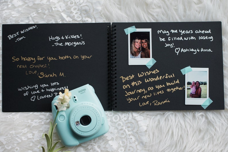 May include: A black scrapbook with gold lettering and handwritten messages. The scrapbook is open to reveal photos and messages. The scrapbook is on a white fuzzy surface with a light blue instant camera.