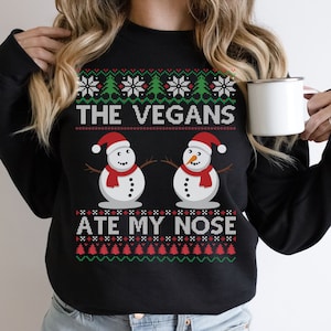 Op de afbeelding: Zwart sweatshirt met een feestelijk ontwerp. De tekst "THE VEGANS ATE MY NOSE" staat boven twee sneeuwmannen met rode sjaals en kerstmutsen. Het ontwerp bevat sneeuwvlokken en kerstbomen.