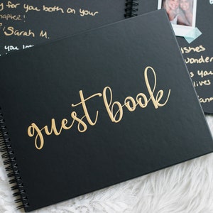 May include: A black guest book with gold lettering that reads "guest book". The book is closed and has a spiral binding.
