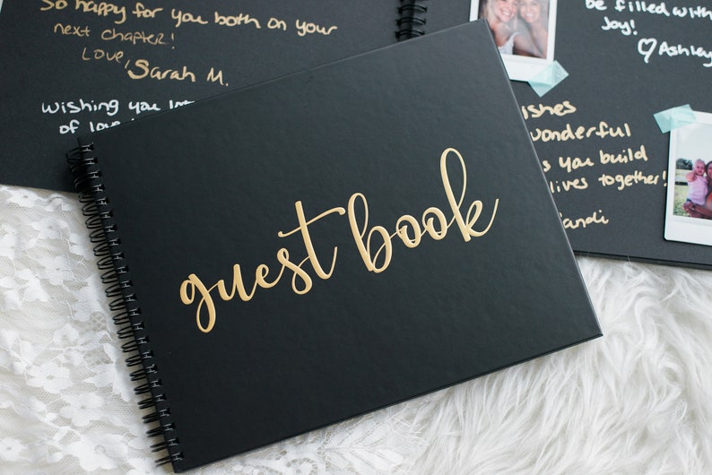 May include: A black guest book with gold lettering that reads "guest book". The book is closed and has a spiral binding.