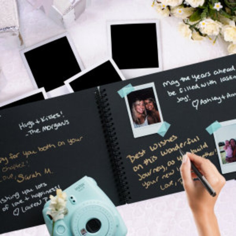 May include: Black guest book with a spiral binding. The book is open to a page with handwritten messages and a few Polaroid photos attached with tape. The text reads "Hugs & Kisses! -The Morgan's" and "May the years ahead be filled with lasting joy! Ashley + Am".