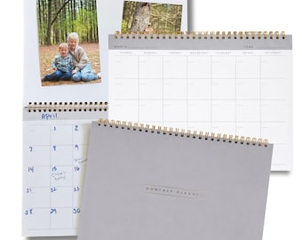 May include: A collection of spiral-bound calendars and planners. One calendar displays a photo and the month of April. Another calendar shows a weekly layout. A gray planner with "MONTHLY PLANNER" printed on the cover is also visible.