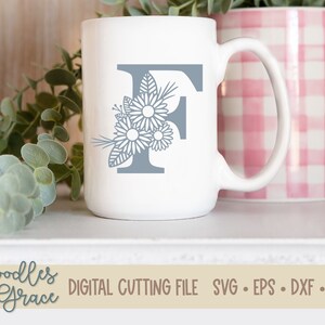 May include: A white ceramic mug with a gray letter F and floral design. The mug is sitting on a white surface with a pink and white checkered mug behind it.