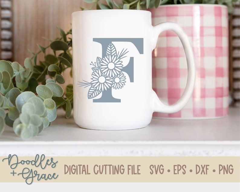 May include: A white ceramic mug with a gray letter F and floral design. The mug is sitting on a white surface with a pink and white checkered mug behind it.