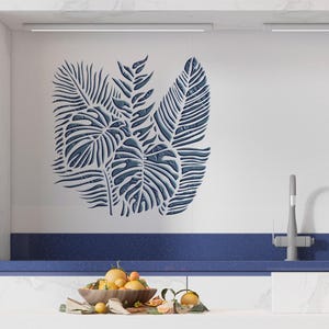 Pode incluir: Um adesivo de parede com um grupo de folhas tropicais azul-escuras, incluindo formas de monstera e samambaia, com uma aparência texturizada. O design é aplicado em uma superfície branca, exibindo recortes intrincados e padrões botânicos.