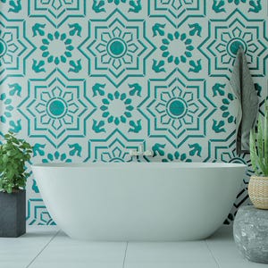 May include: A white freestanding bathtub positioned against a wall featuring a repeating teal and white geometric star pattern. A green plant in a dark rectangular planter sits on the left, and a potted plant sits on a grey stone stool on the right.