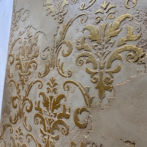 May include: A decorative wall panel featuring a textured beige plaster surface with a raised, metallic gold damask pattern. The finish displays a distressed, uneven appearance with visible cracks and varying depths across the surface.