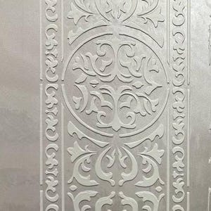 Könnte beinhalten: Nahaufnahme einer Wand mit strukturiertem, hellgrauem Hintergrund. Die Wand weist ein sich wiederholendes Muster aus weißen, verzierten Blumen- und Scroll-Designs auf. Ein vertikaler Rand mit einem kreisförmigen Design befindet sich links.