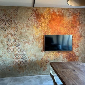 Op de afbeelding: Een muur met een getextureerde, verweerde afwerking in beige, roest- en oranjetinten, bedekt met complexe geometrische patronen. Een flatscreen-tv is centraal op de muur gemonteerd, boven een houten tafel en stoelen.