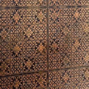 May include: Close-up of a decorative, textured panel with a repeating pattern. The design features squares with ornate, embossed floral and geometric elements in shades of bronze and black. The panel has a vintage or antique aesthetic.