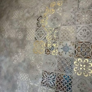 Pode incluir: Uma parede com um fundo cinza texturizado com um padrão repetitivo de ladrilhos quadrados. Os ladrilhos são decorados com vários desenhos florais e geométricos em tons de branco e dourado, criando um efeito decorativo e artístico.