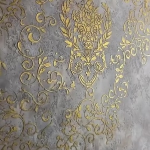 Puede incluir: Una pared gris texturizada con un patrón floral dorado. El diseño presenta remolinos ornamentados, hojas y un motivo floral central, creando una estética clásica y elegante. Los elementos dorados proporcionan un contraste llamativo con el fondo gris.