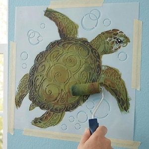 May include: A stencil of a sea turtle with a roller applying green and brown paint. The turtle design includes intricate details and is taped to a light blue wall. Bubbles are stenciled around the turtle.