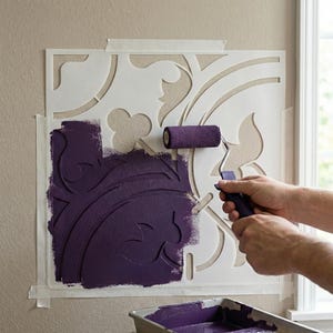 Op de afbeelding: Een decoratieve muursjabloon met een bloemenontwerp wordt geverfd met een paarse verfroller. De sjabloon is op een beige muur geplakt en een bakje paarse verf is zichtbaar. Een doe-het-zelf project.