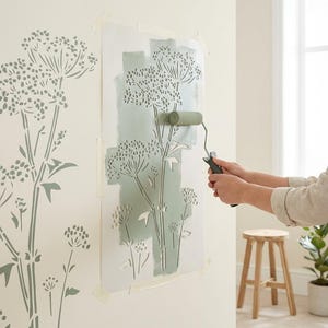 Può includere: Uno stencil floreale viene applicato a un muro con un rullo di vernice verde. Il muro presenta già un motivo floreale a stencil. L'immagine raffigura un progetto di decorazione domestica.