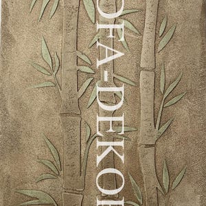 May include: A rectangular wall plaque with a textured tan surface featuring a relief design of three bamboo stalks in a light green hue. The vertical text DFA-DEKOR is printed in white in the center, overlaying the bamboo stalks.