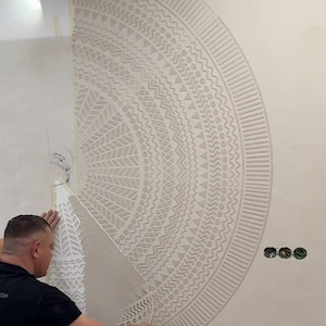 Op de afbeelding: Een persoon die een grote, halfronde muursjabloon aanbrengt met een zich herhalend geometrisch patroon. De sjabloon wordt aangebracht op een lichtgekleurde muur. Het ontwerp bevat driehoeken, zigzags en andere vormen.