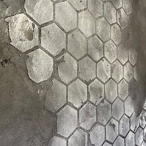May include: A gray concrete surface featuring a repeating hexagonal pattern. The design is applied in a lighter shade of gray, creating a contrast against the darker, textured, and weathered concrete base. The hexagonal shapes vary in clarity.