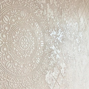 May include: Close-up of a textured wall with a detailed, circular, floral pattern on the left side. The wall has a distressed, off-white color with a slightly rough texture, creating a vintage or antique aesthetic.