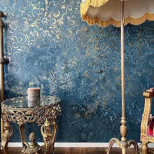 May include: A room with a teal textured wall featuring gold floral patterns. A gold-colored floor lamp with a fringed lampshade stands next to a small, ornate gold table with a glass top. A candle sits on the table.