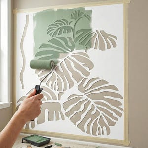 Può includere: Una persona usa un rullo per applicare vernice verde a uno stencil da parete con foglie tropicali. Lo stencil è fissato al muro con del nastro adesivo e il disegno è parzialmente dipinto di verde chiaro. Sono visibili una vaschetta per la vernice e altri materiali per la pittura.