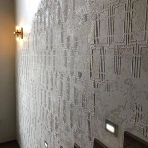 May include: A textured, light gray wall with a repeating geometric pattern. The wall is illuminated by a warm-toned sconce and recessed lighting. Wooden stairs are visible in the foreground, suggesting an interior design theme.