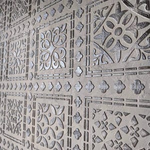 May include: A textured wall featuring a repeating pattern of silver floral and geometric designs. The pattern is embossed on a light beige background, creating a three-dimensional effect. The design is reminiscent of vintage tiles.