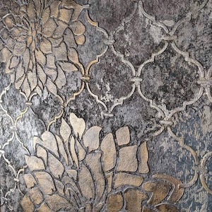 May include: Close-up of a textured wallpaper with a floral and geometric pattern. The design features large, stylized flowers in shades of gold and bronze, set against a distressed gray and blue background. The pattern includes a repeating quatrefoil motif.