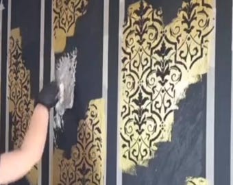 May include: A person applying gold paint with a trowel to a black wall, using a stencil to create a decorative pattern. The pattern is a repeating floral design. White tape frames the sections being painted.