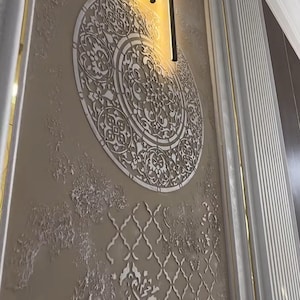 Puede incluir: Una pared con un acabado beige texturizado, con un elemento decorativo blanco, circular y grande con un patrón intrincado. Una lámpara dorada está montada arriba. La pared también tiene molduras blancas con detalles dorados.