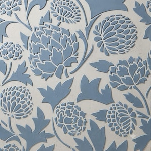 Pode incluir: Um painel de parede quadrado com um padrão floral tridimensional em relevo de flores e folhas azuis semelhantes a cardos sobre um fundo texturizado branco sujo. O design consiste em múltiplas formas botânicas estilizadas.