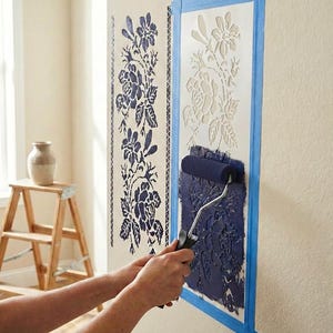 Puede incluir: Una persona usa un rodillo para aplicar pintura azul oscuro a través de una plantilla floral en una pared beige. La plantilla está pegada a la pared con cinta adhesiva azul. Un patrón floral completo es visible a la izquierda. Un taburete de madera y un jarrón están en el fondo.