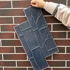 May include: Two dark gray, textured, rectangular tiles with a stone-like appearance are held against a brick wall. The tiles have a pattern of rectangular shapes and lines, creating a faux stone effect. The brick wall is a mix of brown and red bricks.