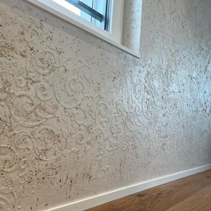 May include: A textured wall with a cream-colored base and a raised, swirling floral pattern. The wall has a distressed appearance with brown and gray speckles. A white window frame and baseboard are visible.