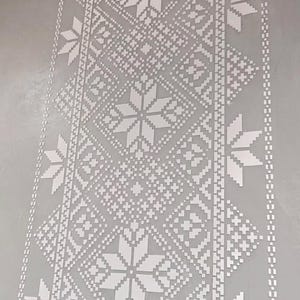 Może przedstawiać: Szara ściana z powtarzającym się białym wzorem geometrycznym. Wzór zawiera kształty przypominające gwiazdy i wzory rombów, tworząc symetryczny, teksturowany wygląd. Wzór jest zorientowany pionowo, odpowiedni do tapet lub zastosowań dekoracyjnych.