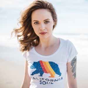May include: A woman with long brown hair is wearing a white t-shirt with a blue bear graphic and the text "CALIFORNIA SOUL".