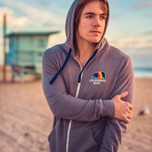 May include: A grey zip-up hoodie with a navy blue drawstring and a graphic of a bear with the text "California Soul".