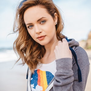May include: A woman with long, wavy brown hair is wearing a grey hoodie with a blue zip and a white t-shirt with a rainbow graphic and the text "California Soul".