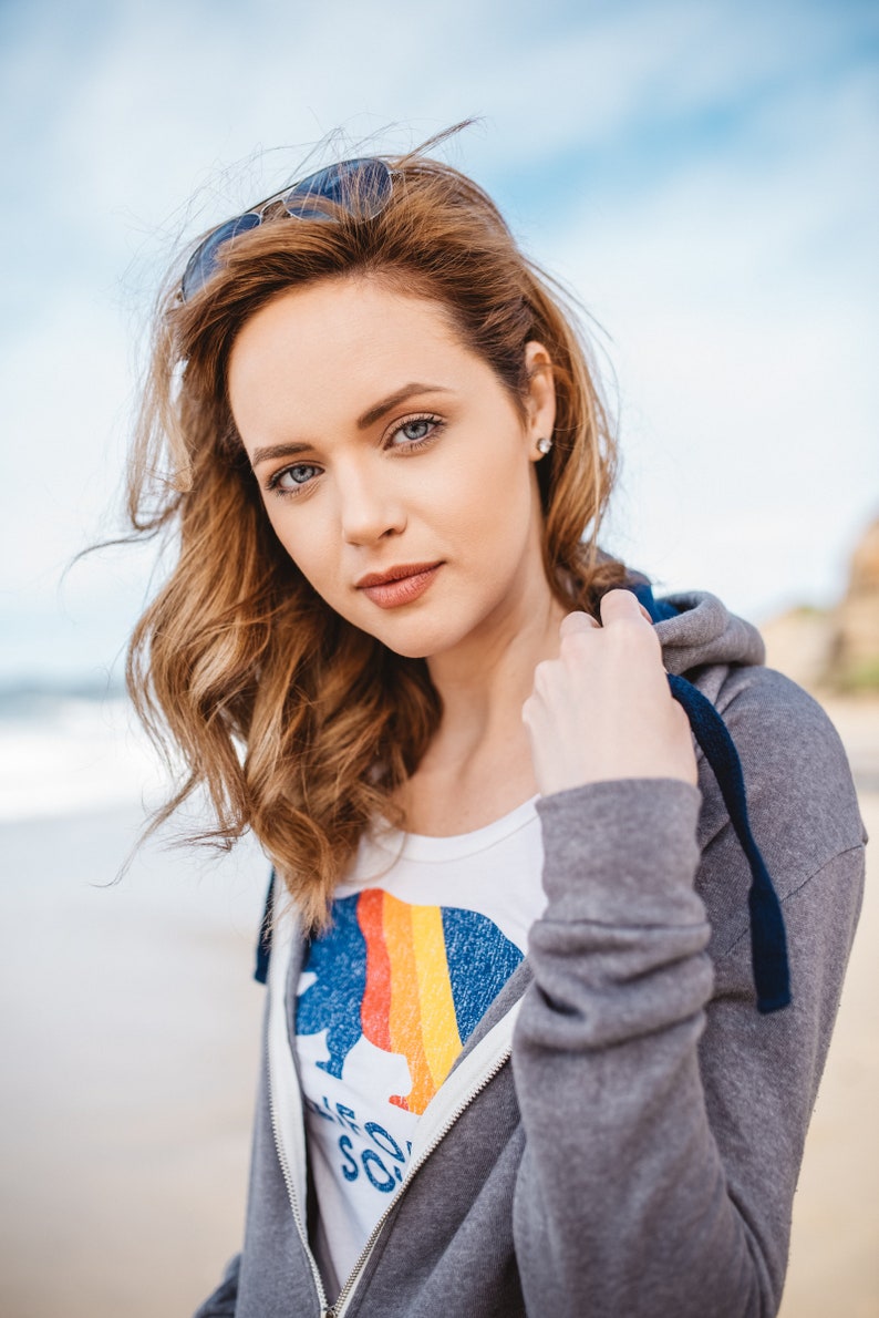 May include: A woman with long, wavy brown hair is wearing a grey hoodie with a blue zip and a white t-shirt with a rainbow graphic and the text "California Soul".