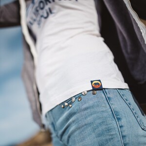 May include: A white t-shirt with the text "California Soul" printed on the front. The shirt has a blue and yellow patch with a bear silhouette on the back. The shirt is tucked into blue jeans with silver studs.
