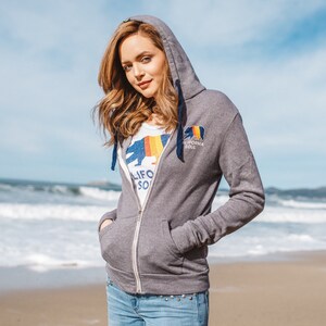 May include: A grey zip-up hoodie with a California bear logo and the text "California Soul". The model is wearing the hoodie with the zipper pulled up and is standing on a beach.